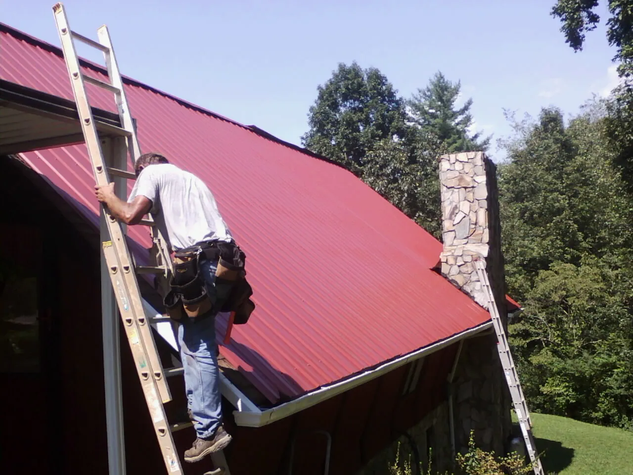 Roofer on a ladder beside a red metal roof and stone chimney — Soffit & Fascia Repair by Roofing Brothers in Weatherford